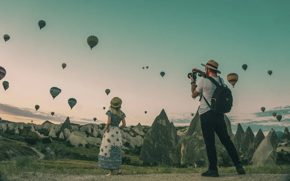 Two travelers watch and photograph hot air balloons over Cappadocia, Türkiye